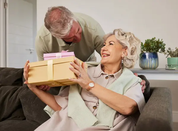 惊喜和一对老夫妇在他们的家客厅庆祝生日在一起 礼物和结婚纪念日 丈夫坐在沙发上给妻子一个盒子 — 图库照片