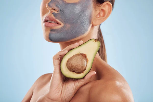 Woman, avocado and face mask for skincare, natural beauty and vitamin d benefits on studio, blue background. Person or model with charcoal facial, green fruits and healthy skin care for dermatology.