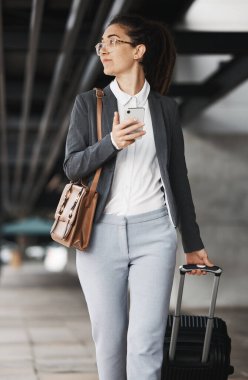 Şehirdeki bir havaalanı otoparkında yürüyen bir iş kadınıyla telefon, görüş ve bavul. Cep telefonu, bavul ve uluslararası bir iş gezisinde genç bir bayan profesyonelle düşünme.