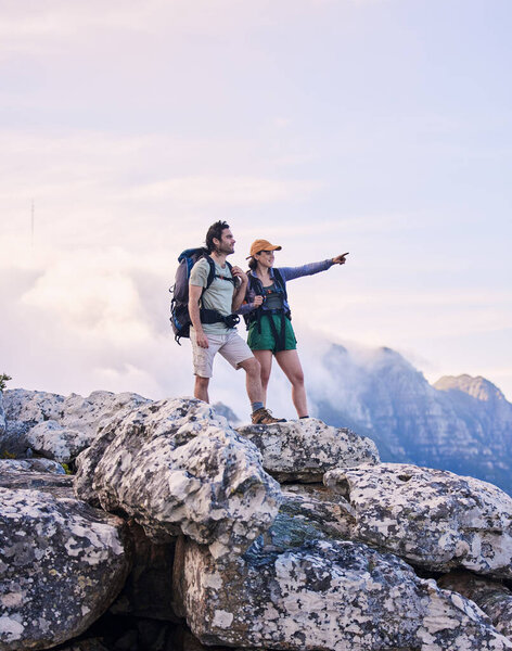 Mountains, hiking and pointing, man and woman on peak for adventure in nature, landscape and travel. Outdoor trekking, couple on cliff and relax in scenic clouds for natural journey, view and looking.