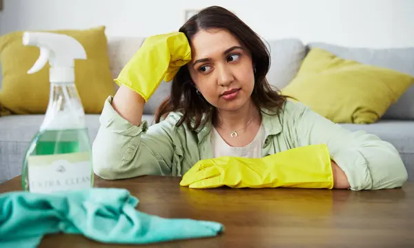 Woman, table and thinking for cleaning home, stress and tired with gloves, cloth and chemical product. Girl, ideas and fatigue with spray, bacteria or sitting in living room with anxiety for hygiene.