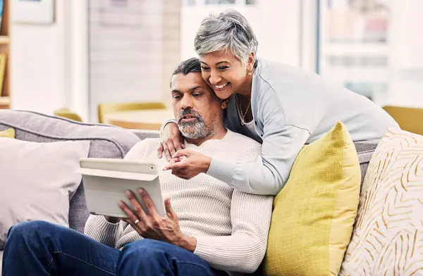 Love, hug and mature couple on a tablet networking on social media, mobile app or the internet. Happy, embrace and senior man and woman browsing on digital technology on a sofa in living room at home.
