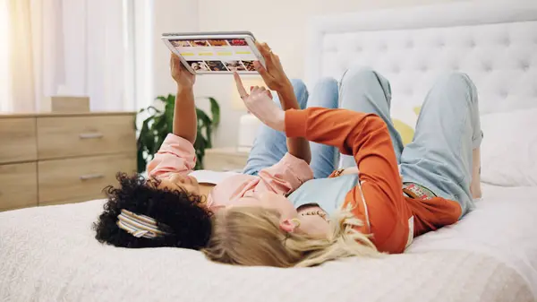 Friends relax with tablet to scroll on website for a movie or film choice lying on bed together at home. Technology, decision or people on internet search for ecommerce online shopping on app screen.
