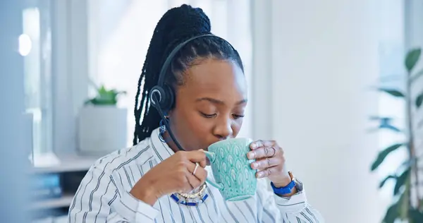 Happy black woman, call center and drinking coffee in customer service, support or telemarketing at office. African female person, consultant or agent enjoying hot beverage in contact us at workplace.