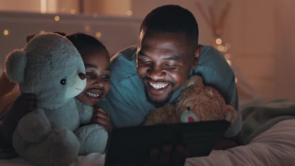 ベッド 夜と少女と父親 タブレットと幸せ漫画 家とリラックスして接続 テクノロジー テディベア インターネットを持つ子供 子供を持つブラックファミリー 親とパパ — ストック動画