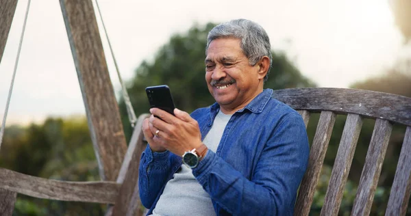 Senior man, happy and smartphone on bench for communication, connection and social media in park. Mature pensioner, smile and technology for lounge in nature, digital games and wellness on retirement.