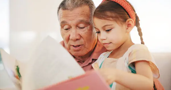 Grandparent, kid and reading book with smile, home and child development, grandchild and couch. Man, lounge or bonding together for relationship, family and retired with girl, house and happy.