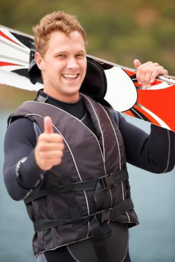 Spor, baş parmaklar ve gölün kenarında sörf, egzersiz ve eğlence hobisi için sörf tahtası olan bir adamın portresi. Fitness, emoji el işareti ve su kayağı için tahtası olan kişi özgürlük, macera ve eğlence için.