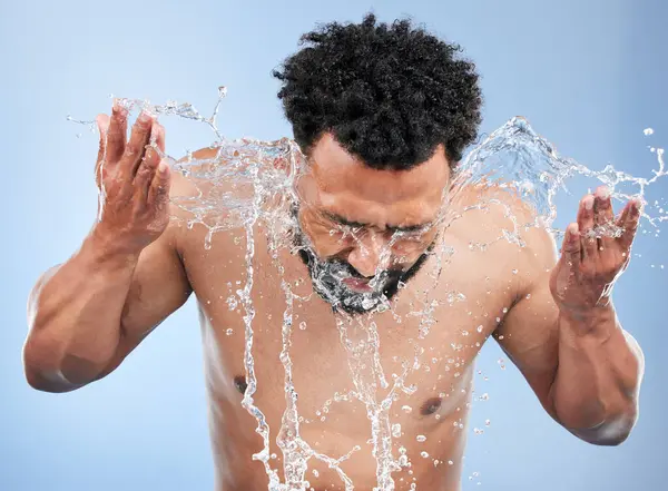 Black man, water splash and face wash for skincare, hygiene or grooming against a blue studio background. African male person, model and aqua for body hydration, shower or facial skin treatment.