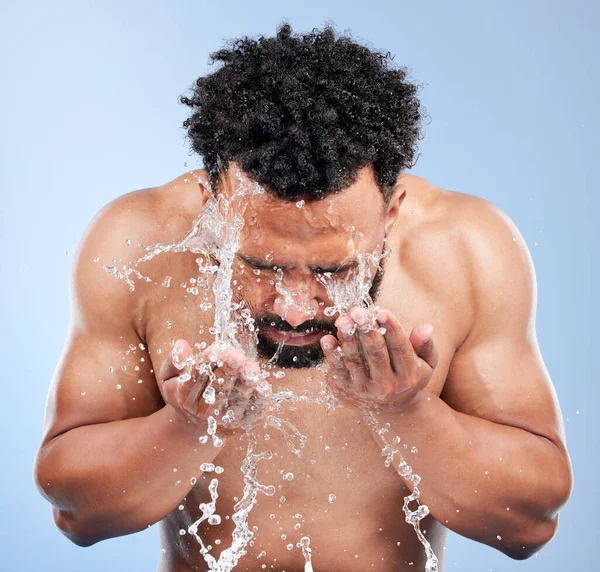 Black man, water and washing face for skincare, hygiene or grooming against a blue studio background. African male person, model and aqua splash for body hydration, shower or facial skin treatment.