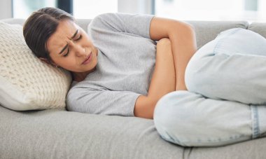 Woman, stomach pain and stress on sofa, sick or menstruation with anxiety, gas or virus in home living room. Girl, abdomen and emergency for gut health, constipation or cramp on lounge couch in house. clipart
