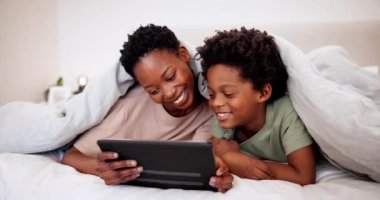 Anne, yatak odasında tabletteki çocukla gülümse, çizgi film, film ve videoyu battaniyeyle birlikte öğren ve izle. Afrikalı çocuk, teknisyen ve anne yatakta, internet uygulaması ve aile içi rahatlama hakkında konuşuyorlar..