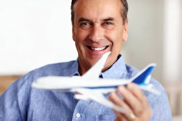 Mature man, portrait and toy plane for travel, vacation and holiday planning with a smile at home. Happy, airplane model and retirement of a male person with confidence from trip and air traveling.