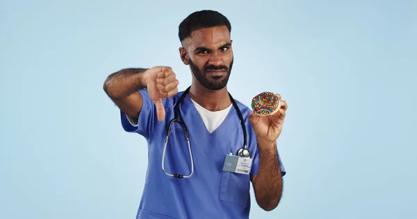 Man, portrait and doctor with thumbs down, donut or bad review for diet against a blue studio background. Brazilian person or medical nurse rejection, no emoji or sign for chocolate dessert on mockup.