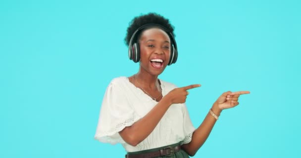 ヘッドフォン付きのポインティング 黒人女性 ブルーバックグラウンドのモックアップスペースに隔離されたスタジオで幸せでダンス ラジオ ストリーミング ヒアリングプロモーション フェイスポート — ストック動画