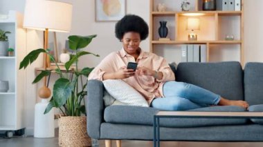 Afro kadın telefonda gülüyor, abonelik film kanalında komik komedi videoları izliyor. Gülümseyen, mutlu bir kadın. Oturma odasındaki kanepede oturuyor. Rahatlıyor ve sosyal medyada geziniyor..