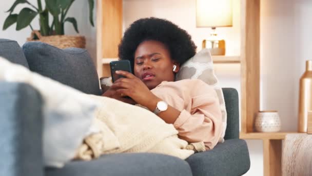 疲惫疲惫的女人在沙发上休息 在家用手机打字 在一个无聊的周末 一个年轻的非洲女性在家里的沙发上放松和打呵欠 一个昏昏欲睡的女人在社交媒体上打滚 — 图库视频影像