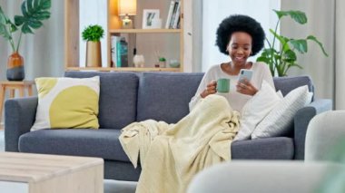Mutlu kadın evde bir kanepede kahve içerken telefonda sosyal medya haberleri okuyor. Genç Afrikalı Amerikalı kadın internette ilginç komik içerikler yazıyor ve paylaşıyor..