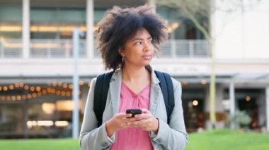 Telefonla yazı yazan ve üniversite kampüsünün dışında yürüyen mutlu bir kız öğrenci. Kablosuz bir cihazda sosyal medyada gezinirken afro saçı ve sırt çantasıyla gülen şık bir üniversite öğrencisi..