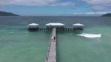 Bir çift rıhtımda yürüyor, okyanustan drona ve su üzerindeki kulübeye tropik tatilde dalgalar, seyahat ve özgürlükle birlikte. Kumsal, tatil ve dinlenme, ahşap köprüdeki insanlar gökyüzü ve hava manzaralı