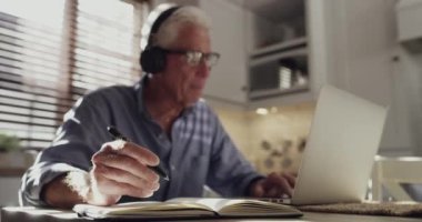 Ev bilgisayarı, defter ve iş bilgileri yazan yaşlı bir adam. Mutfaktaki online raporun özeti. Kulaklıklar, günlük ve araştırma yapan yaşlı kişi, uzaktan çalışma ve planlama gündemi.