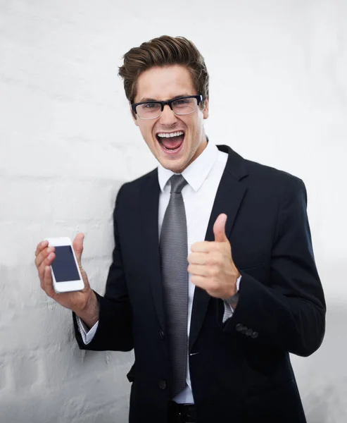 Excited businessman, portrait and phone with thumbs up for success against a gray wall background. Happy man or employee smile with mobile smartphone, like emoji or yes sign for winning or good job.