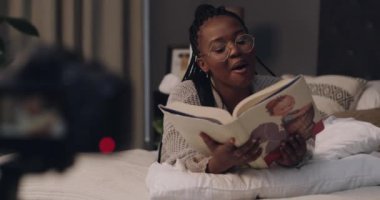 Genç kadın, sosyal medya kanalı için çevrimiçi kayıt ile yatakta video kaydetmek için kitap okuyor. Siyah insan, mutlu ve online podcast için canlı yayın ve yatak odasında peri masallarının etkisinde..