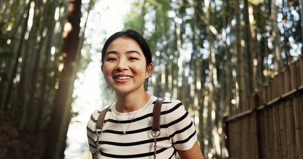 Japanese woman, portrait and bamboo in forest with smile, pride and backpack for travel on holiday in bush. Girl, person and happy with hiking by plants, freedom and outdoor by trees, woods or nature.