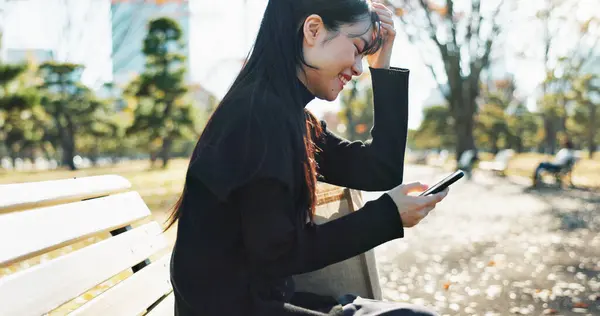 Japanese woman, bench and smartphone in park with relax, happiness and social media in sunshine in city. Female person, sitting and wellness with cellphone in town, streaming and digital technology.