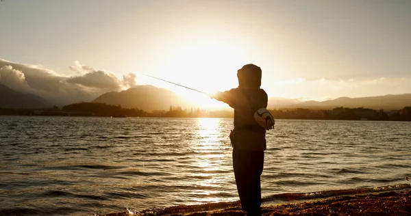 Person, fishing and water in morning, sunrise and sport by ocean, lake and river with equipment in nature. Fisherman, line and catch fish for games, food and silhouette in sunshine, dawn and horizon.