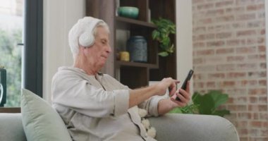 Kulaklıklar, ev ve cep telefonu olan yaşlı bir adam, dinlenme ve oturma odasında müzik dinleme. Bir dinlenme odasında emekli, apartman dairesi ve akıllı telefonu, sesi ve uygulaması olan yaşlı bir adam..