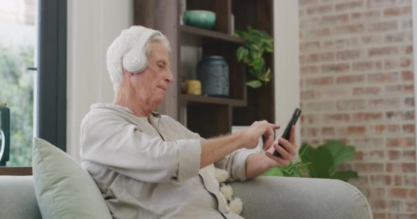 家里的耳机和老年人带着手机 在客厅里放轻松 放音乐 养恤金领取者在休息室 公寓和老年人与智能手机 收音机和音频与声音和应用程序 — 图库视频影像