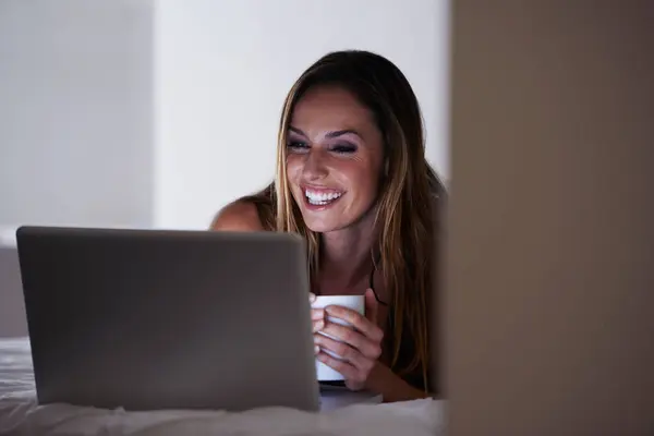 Happy woman, laptop and coffee on bed in morning for online entertainment or streaming at home. Young female person smile with beverage, latte or tea in relax on computer for social media in bedroom.