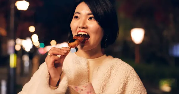 Woman, street food and eating Japanese snack for travel experience, hungry or local trip. Female person, sidewalk and night or bite grilled mochi on road for vacation culture, adventure or tradition.