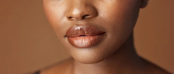 Closeup, lips and black woman with beauty, smile and skincare on a brown studio background. African person, girl and model with gloss, cosmetics and dermatology with clear skin, smooth and aesthetic.