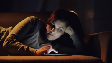 Kadın, tablet ve gece kanepe üzerinde online yayın, sosyal medya veya evde araştırma okumak için. Akşamın geç saatlerinde oturma odasındaki kanepede uzanan ve interneti taramak için teknolojiyi kullanan bir kadın..