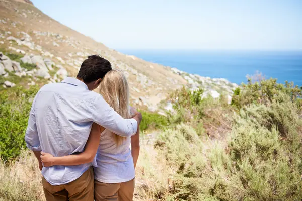 Couple, hug and love on mountains for travel, journey and holiday in nature with blue sky and ocean view. Back of romantic people on eco friendly date, hiking on hill and valentines day or tourism.