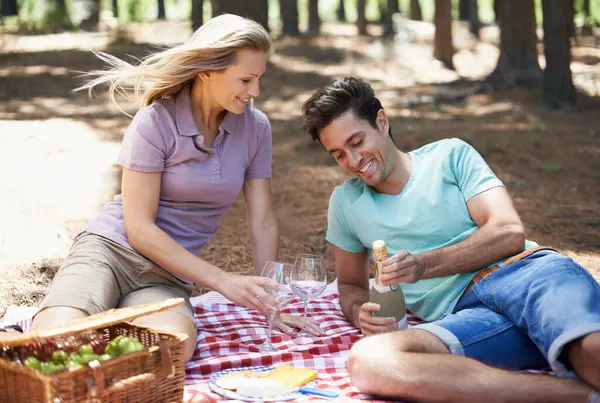Couple, picnic and champagne in nature with love, romantic celebration and summer date on valentines day. Young people with food, fruits and bottle with wine glasses in woods or forest on anniversary.
