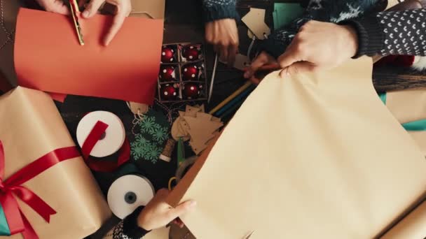 クリスマスのための手 人々およびギフト包装 リボンが付いている芸術および技術 現在のための札かラベルが付いている茶色のペーパーを切って下さい ボックス ホリデーシーズンのパッケージとの友人 創造性のトップビュー — ストック動画