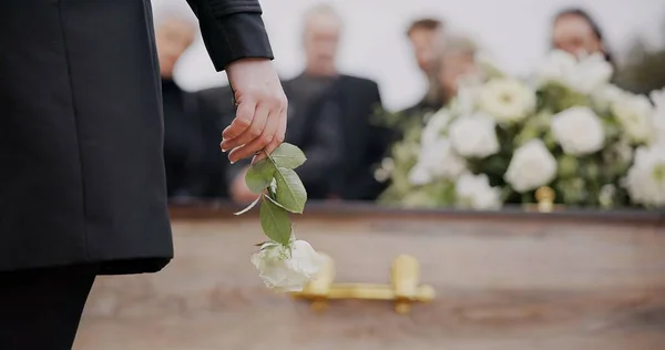 Death, funeral and hand of man with flower at coffin, family at service in graveyard for respect. Roses, loss and people at wood casket in cemetery with memory, grief and sadness at grave for burial