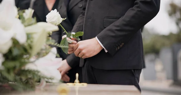 Hand, rose and a person at a funeral in a graveyard in grief while mourning loss at a memorial service. Death, flower and an adult in a suit at a cemetery with a coffin for an outdoor burial closeup.