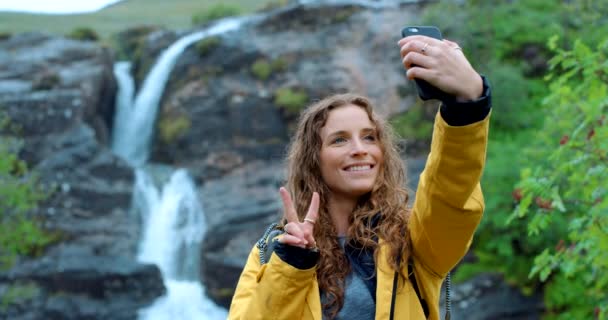 自私自利 徒步旅行 带着和平标志的女性自然旅行博客 探险或度假 徒步旅行 佳节快乐的女孩在山中的直播溪流中 为瀑布中的社交媒体追忆 — 图库视频影像