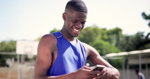 ブラックマン 携帯電話 バスケットボールコートで喜んでメッセージ 屋外でのトレーニングでプレーヤー アフリカのスポーツ選手 携帯電話 またはインターネットのためのスポーツゲームやモバイルアプリの健全性のためのポジティブ — ストック動画