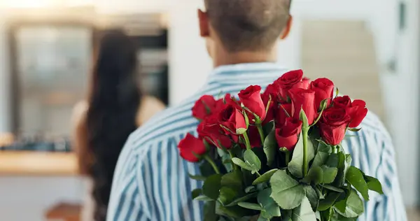Happy couple, red roses and kiss for surprise, anniversary or valentines day in kitchen at home. Face of young man and woman smile with flowers for romantic gift, love or care in celebration at house.