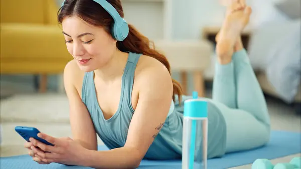 Smartphone, yoga and headphones of woman typing on chat app for fitness communication, social media update or blog writing wellness. Pilates girl with exercise gear and technology in home living room.