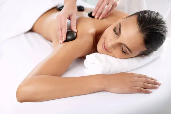 Hot stone, woman and hands massage shoulder for skincare, beauty and pamper body for wellness at luxury salon. Back, rocks and happy person at spa for treatment, relax or health with masseuse.