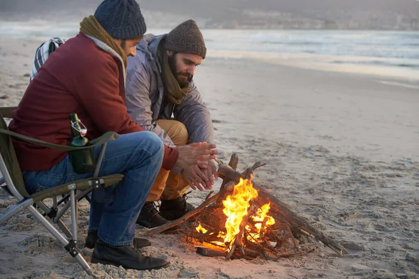 Campfire, friends and adventure with men by the beach at sunset with vacation and camping. Ocean, outdoor and together with travel and people in a trip and journey with fire and conversation by sea.