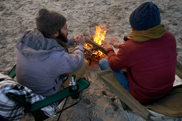 Campfire, friends and talking with men by the beach at sunset with vacation and camping. Ocean, outdoor and together with travel and people back on trip and journey with fire and conversation by sea.