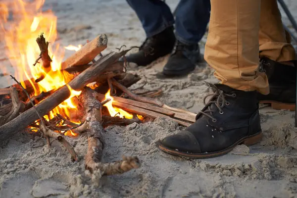 Closeup, campfire and friends at the beach on holiday, vacation and travel to warm up in winter outdoor. Flame, wood and people together by sand at seashore with shoes, adventure and legs in nature.