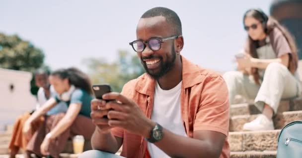 ブラックマン スマートフォン 幸せな学生は ソーシャルメディアでタイピングし ステップで面白いミームを読んでいます キャンパスの屋外でジョーク スクロール モバイルアプリのための大学の電話 — ストック動画